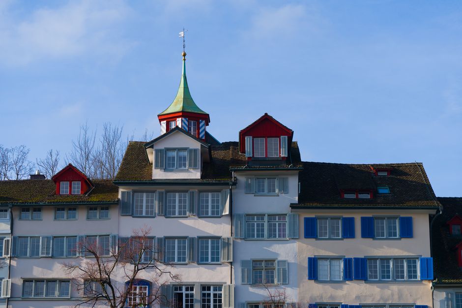 A multi-storey residential building with a white facade and numerous windows, some with blue shutters, situated under a clear blue sky. The roof features several gabled sections with dark tiles and sm